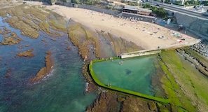 An aerial view of a beach with a rocky shoreline, clear water, and a green barrier separating the beach from the water. -  Aloha Beach House H (Cascais)