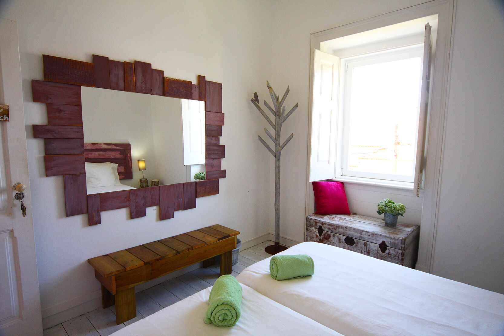 A bedroom with a wooden bench, a large mirror, a bedside table with a plant, and a window with a view.