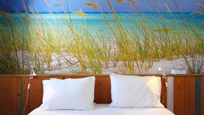 A bedroom with a bed, white pillows, and a view of a beach with tall grass and a clear blue sky. -  Aloha Beach House H (Cascais)