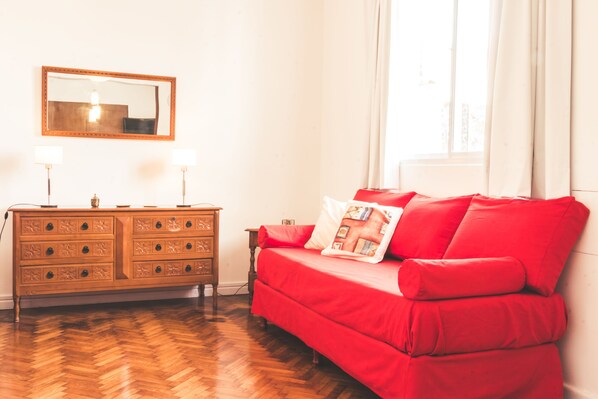 A room with a wooden dresser, a red sofa, a mirror, and a window with curtains. - Apartamento en Av. Belgrano (Buenos Aires)