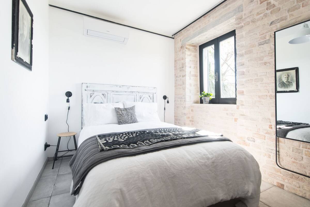 A modern bedroom with a white bed, a brick accent wall, a large window, and a ceiling light.