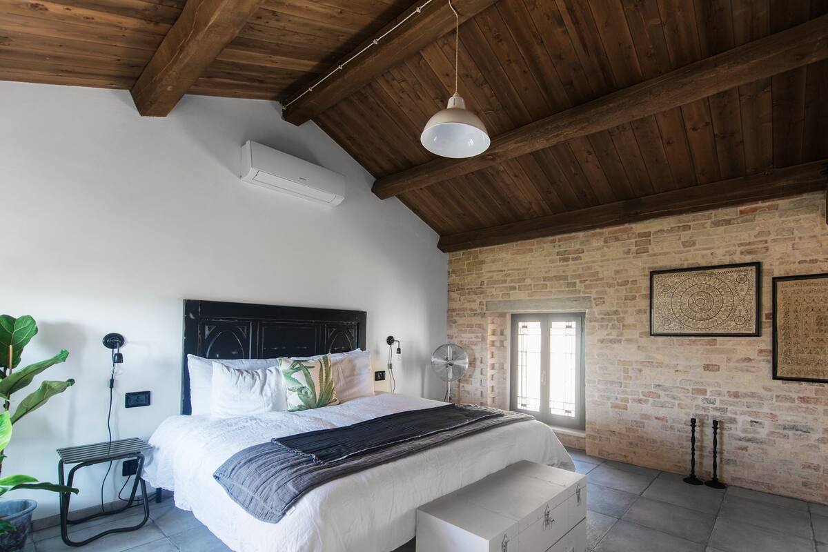 A bedroom with a wooden ceiling, a bed with a white headboard, a bedside table with a lamp, a brick wall, and a window.