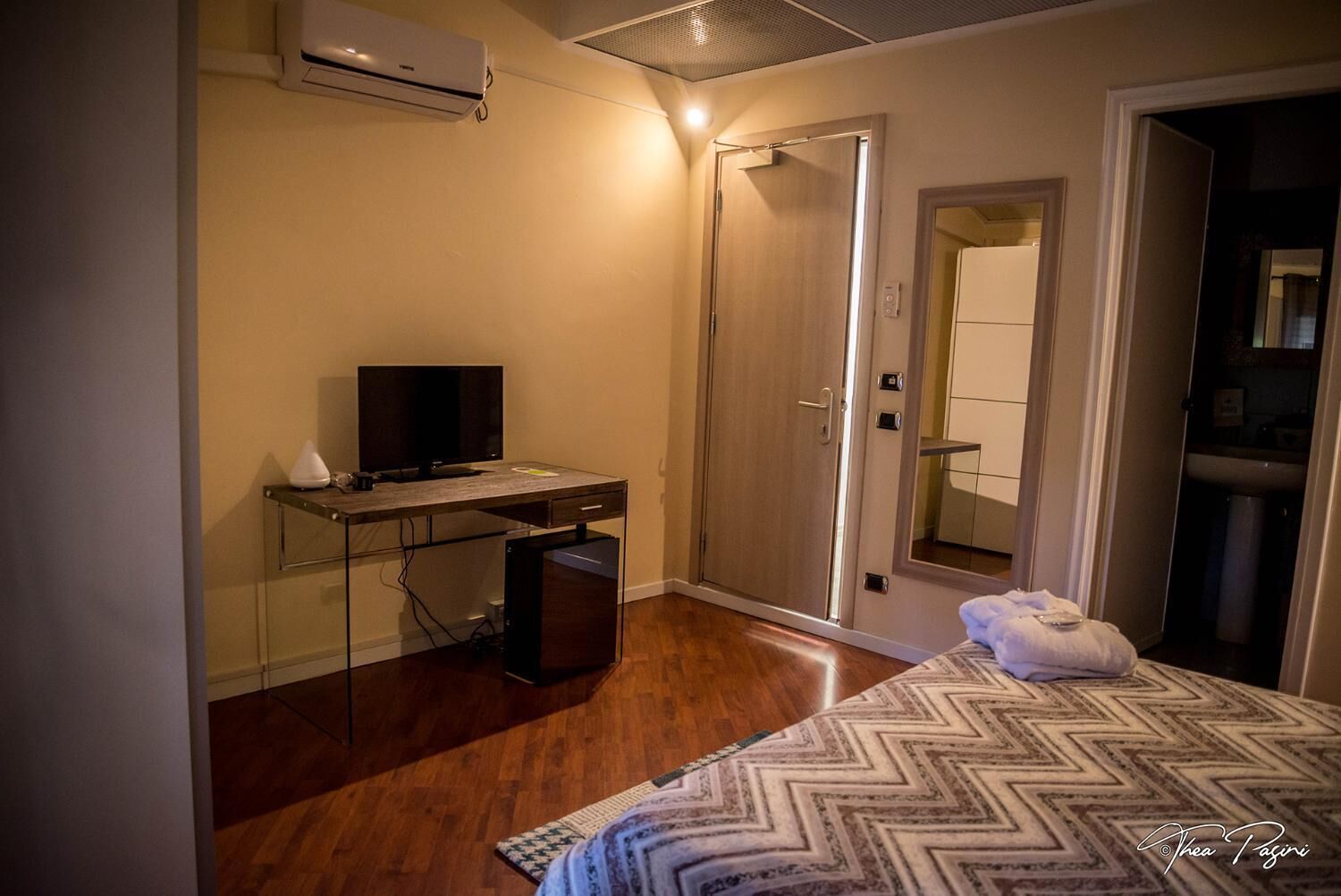 A hotel room with a bed, a desk with a computer, an air conditioner, a door, and a mirror.