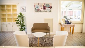 Ein modernes Zimmer mit einem braunen Sofa, zwei weißen Ohrensesseln, einem kleinen weißen Tisch, einer Topfpflanze und einem Blumenbild an der Wand.