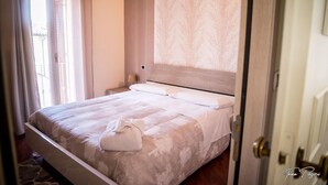 Ein ordentlich bezogenes Bett mit weißer Tagesdecke und Kissen, ein Holz-Kopfteil und ein Fenster mit Vorhängen.