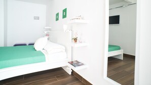 Ein modernes Schlafzimmer mit einer grünen Tagesdecke, weißer Bettwäsche, einem Nachttisch mit Lampe, einer kleinen Ablage mit Büchern und einer Pflanze sowie einem an der Wand befestigten Fernseher.