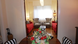 Un comedor con una mesa de madera, sillas y un centro de mesa floral. Hay dos sillones y una mesita con una lámpara.