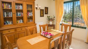 Un coin repas avec une table en bois, des chaises, un meuble contenant des livres, un vase avec des fleurs et une fenêtre avec des rideaux.