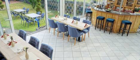A restaurant with a long table and blue chairs, a bar with stools, and a view of a garden through large windows.