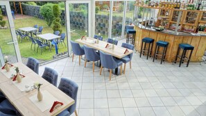 A restaurant with a long table and blue chairs, a bar with stools, and a view of a garden through large windows. - Hotel Opal (Laatzen)