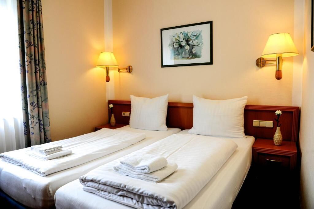 A hotel room with two beds, each with a neatly folded white towel and a small bedside table with a vase.