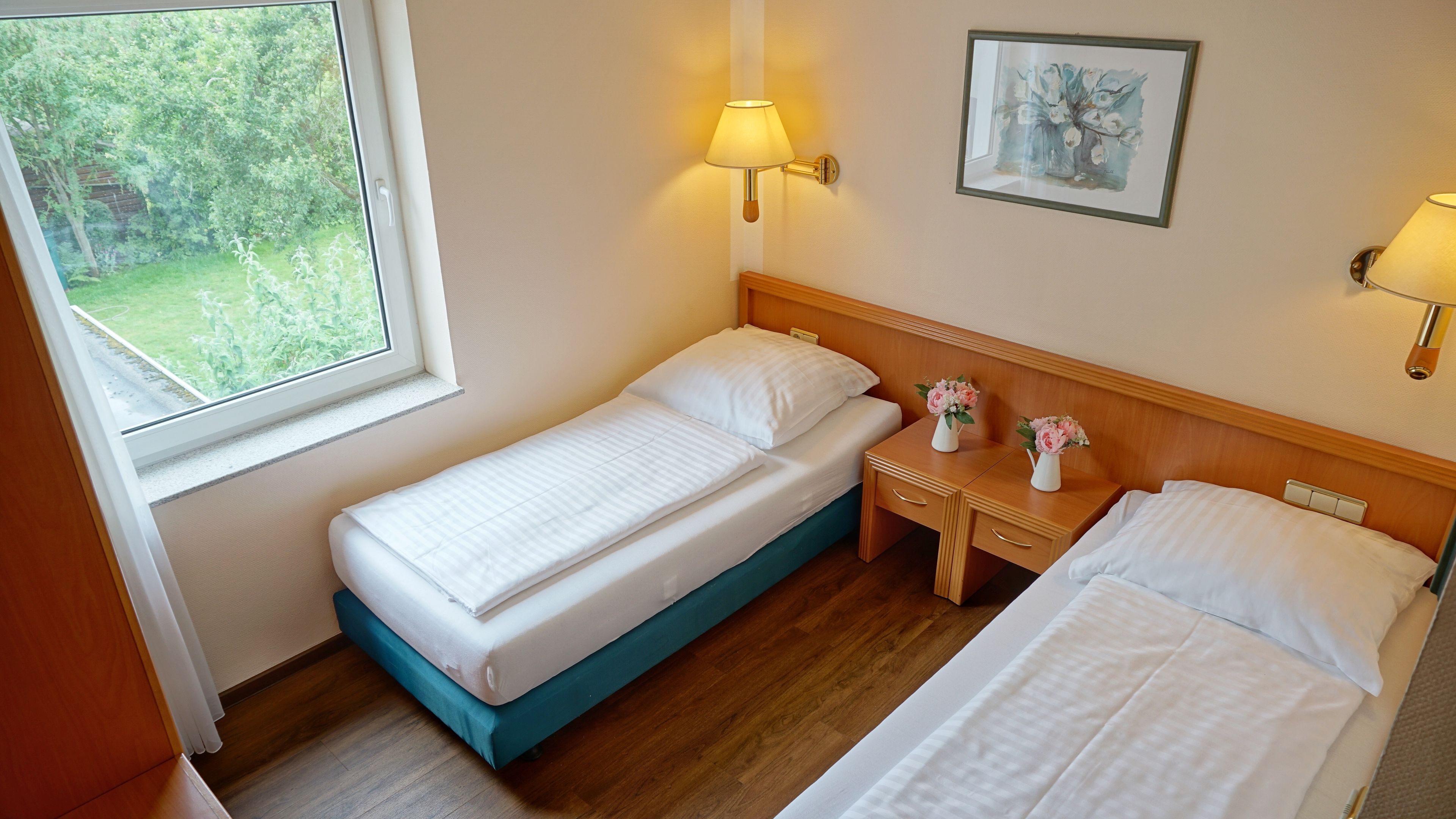 A hotel room with two single beds, a small wooden nightstand, a window with a view of greenery, and two wall-mounted lamps.