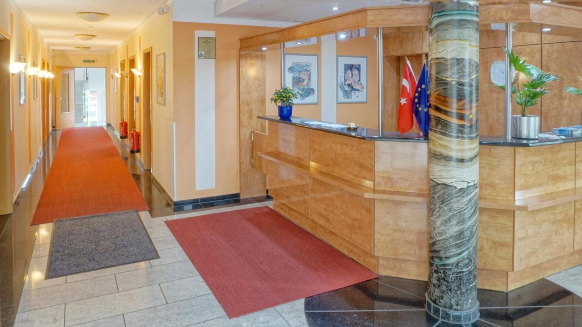 Zona de recepción de un hotel con un mostrador de mármol, alfombra roja y un pasillo con cuadros en las paredes.