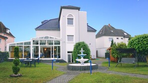 A modern building with a glass entrance, a fountain, and a patio area with tables and chairs. - Hotel Opal (Laatzen)