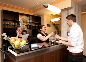 Un mostrador de recepción de hotel con un cuenco de frutas, una planta y una exhibición de vinos al fondo.