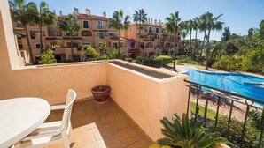 A balcony with a view of a swimming pool, palm trees, and a building with multiple balconies.