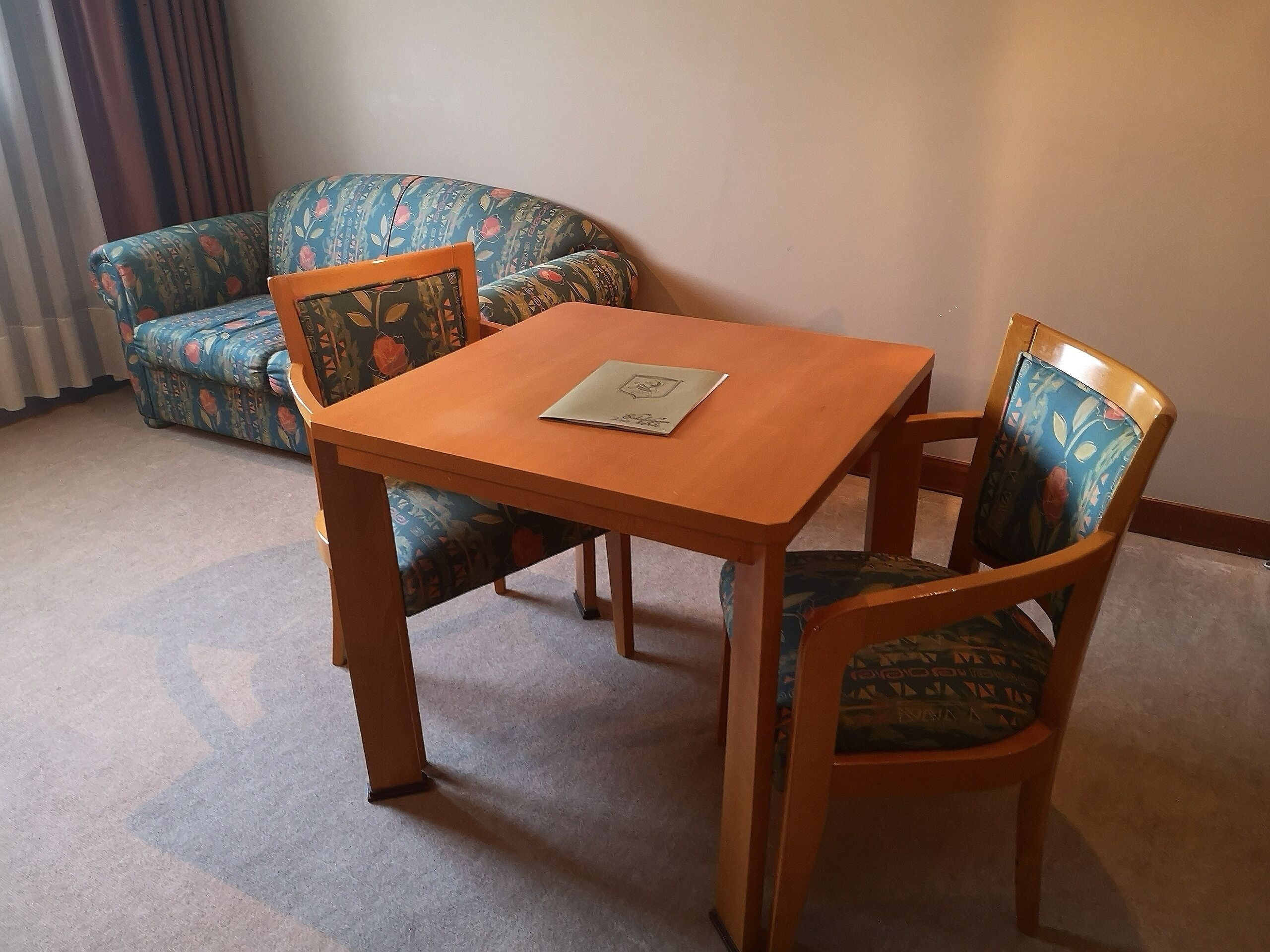 Een kleine kamer met een houten tafel en twee stoelen, een bloemenbank en een radiator.
