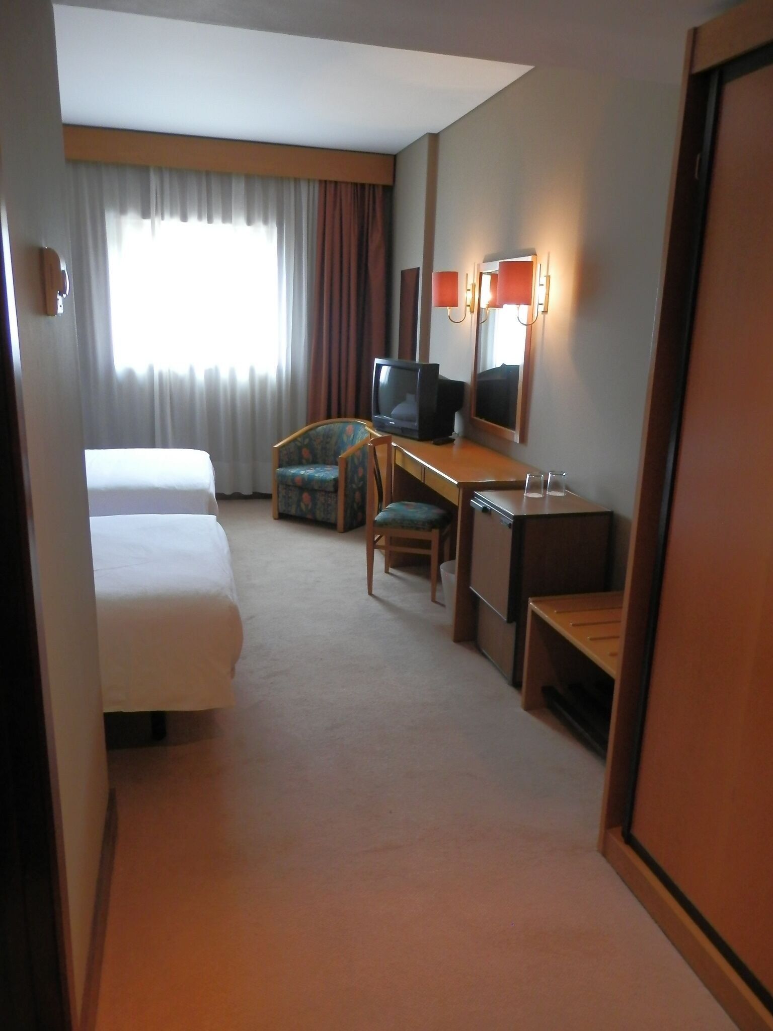 A hotel room with a bed, a desk with a television, a chair, and a small table.