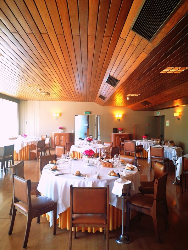 A restaurant with wooden ceiling panels, round tables with white tablecloths, and brown leather chairs. - Hotel Via Norte (Maia)