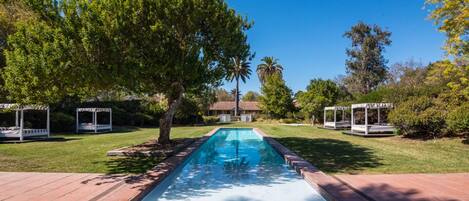 A rectangular swimming pool with a tiled deck, surrounded by grass and trees.