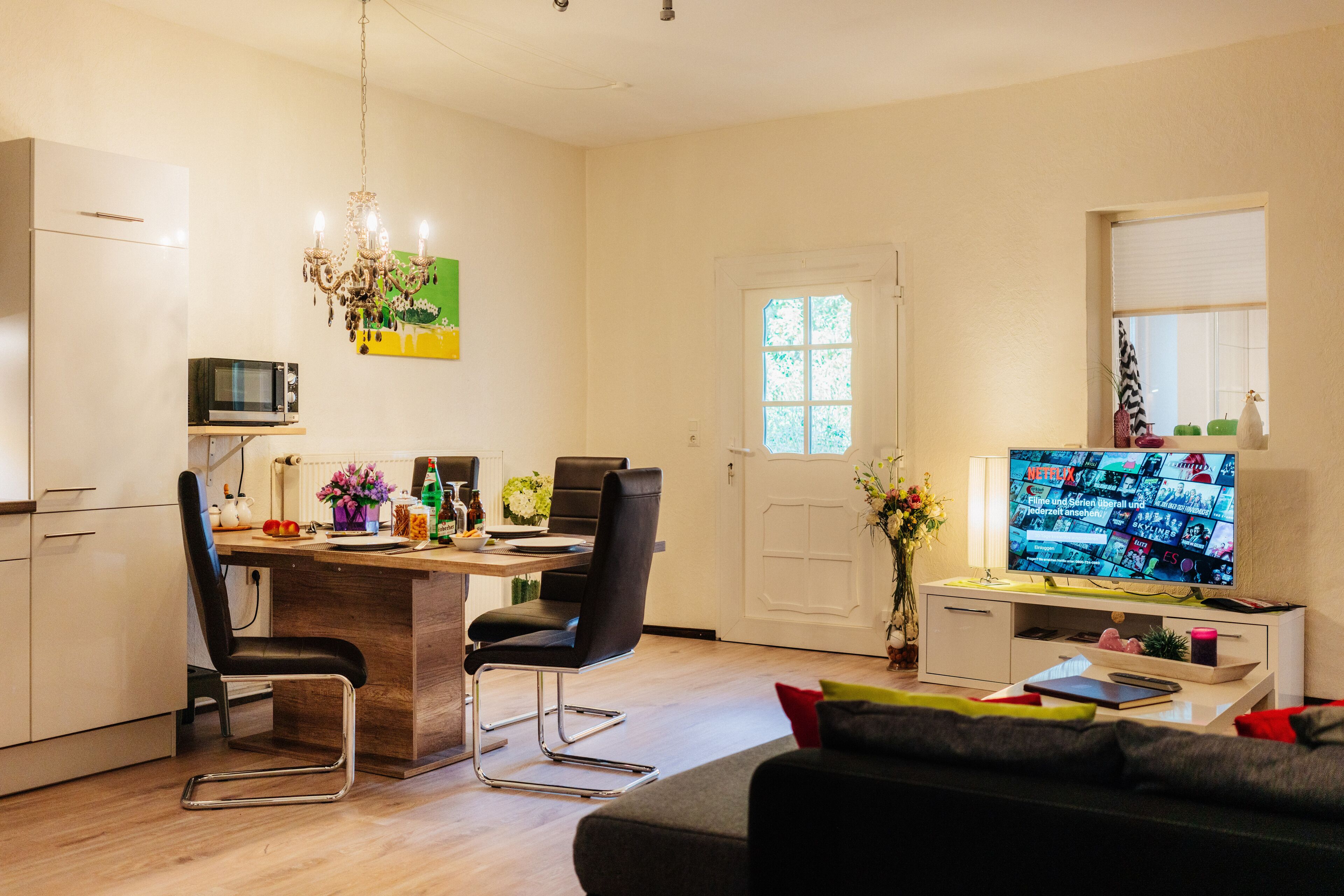 A modern kitchen with a dining area, a living room with a sofa, and a TV displaying a movie.
