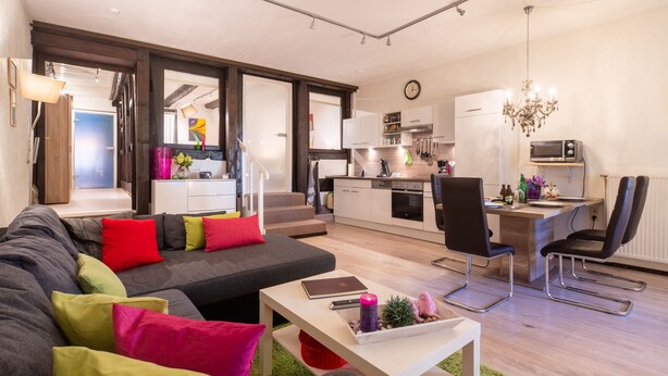 A modern living room with a sofa, a dining area with chairs, a kitchen with appliances, and a chandelier.