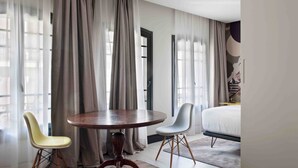 A modern hotel room with a round wooden table, two chairs, a bed, and large windows with sheer curtains. - Amor de Dios 17 Luxury Suites (Madrid)