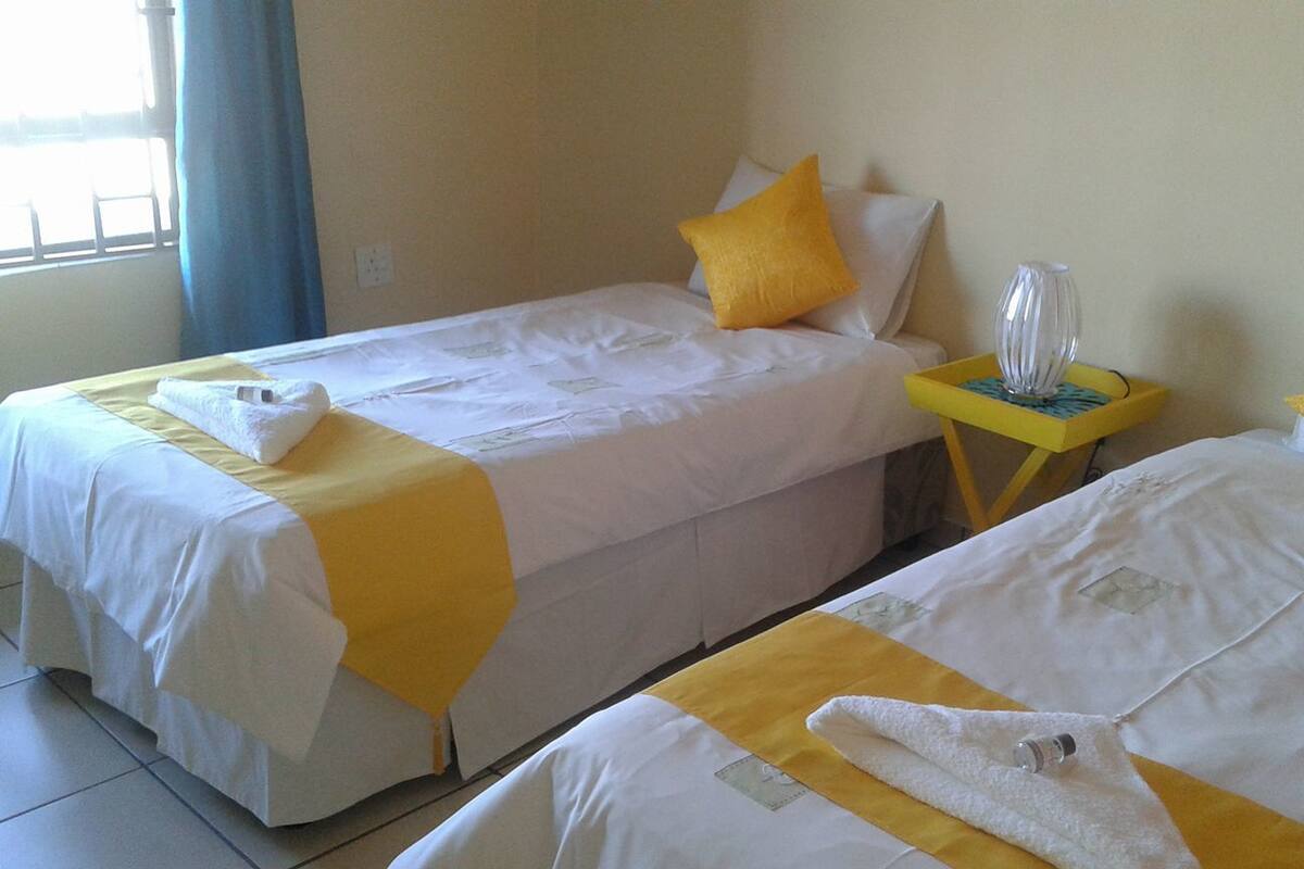 Two single beds with yellow and white bedding, a small table with a vase, and a window with blue curtains.