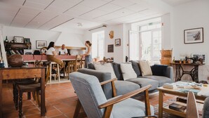 A cozy living area with a sofa, chairs, a coffee table, and a dining table with chairs. There is a kitchen area in the background with a person cooking. - Jaca - Hostel (Machico)