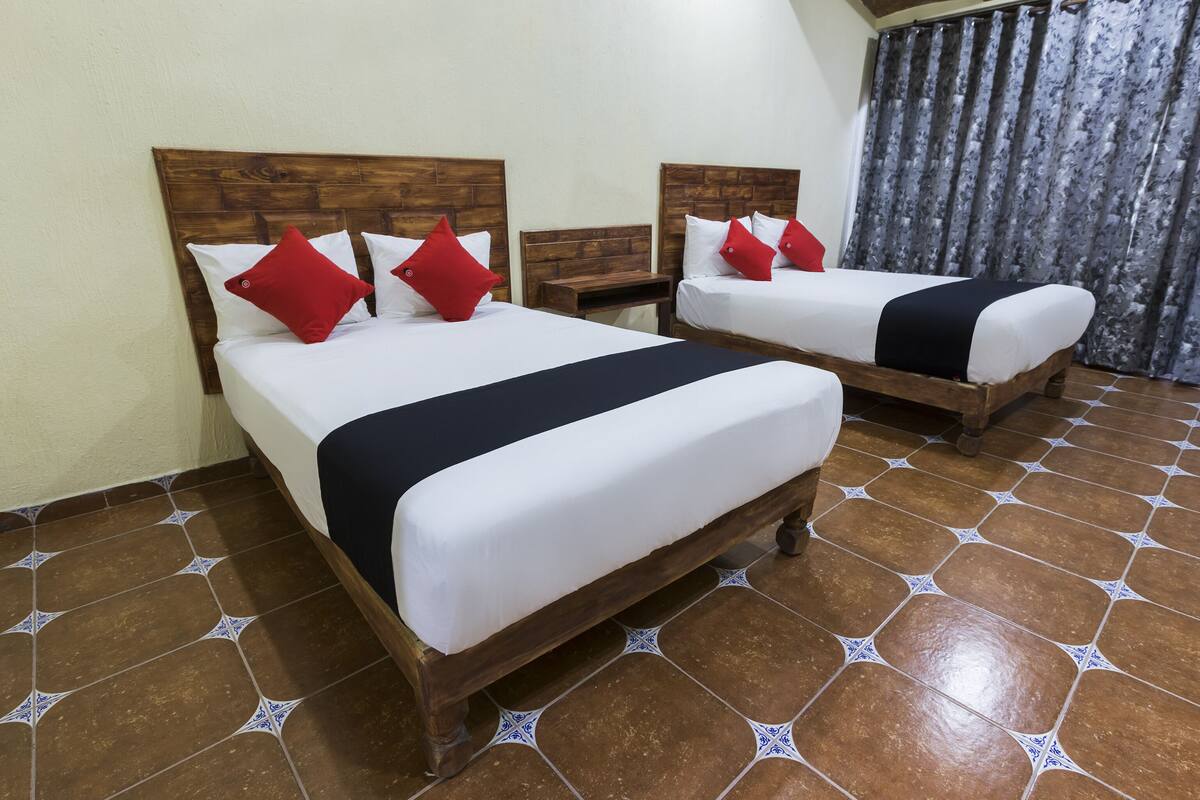 Een hotelkamer met twee houten bedden, elk voorzien van witte beddengoed en rode kussens. De kamer heeft een betegelde vloer met een geometrisch patroon en een houten hoofdbord.