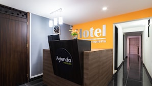 A hotel reception desk with a wooden finish and a black sign that reads "Ayenda HOTELES". There is a clock on the wall above the desk and a vase with flowers on the counter.