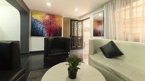A modern living room with a white sofa, black leather armchair, and a round coffee table with a plant. There are two large abstract paintings on the walls and a door leading to another room.