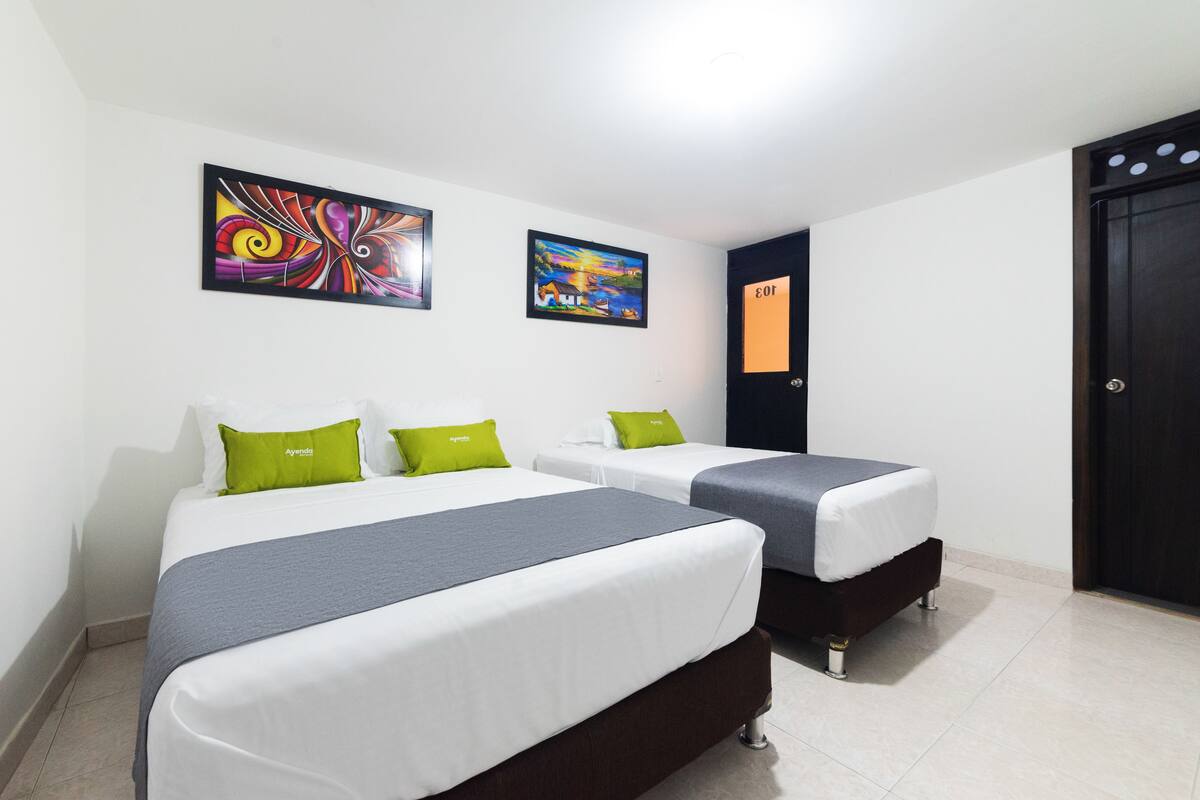 A hotel room with two beds, each with a grey and white duvet, green pillows, and a dark brown headboard. There are two framed artworks on the wall above the beds.