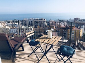 Een terras met uitzicht op zee, met een houten tafel, twee stoelen en een bankje.