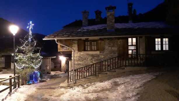Un bâtiment en pierre avec un escalier en bois menant à une porte, un sol recouvert de neige et un sapin de Noël illuminé.