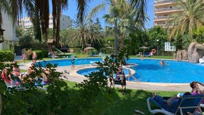 A swimming pool surrounded by palm trees and lounge chairs, with a building in the background.