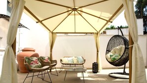Un área de estar al aire libre con techo, una mesa, dos sillas acolchadas, una silla colgante y una cesta decorativa.
