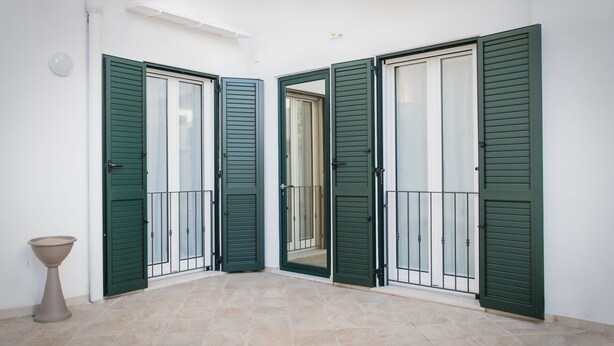 Un pasillo con contraventanas verdes, suelo de baldosas y una maceta decorativa.