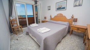 Une chambre à coucher comprenant un lit, une chaise, une table de chevet, une lampe et une vue sur la mer, accessible par une porte coulissante.