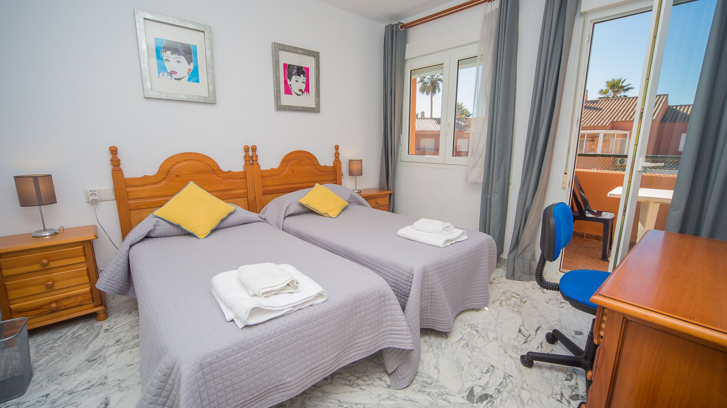A hotel room with two beds, a desk, a chair, and a window with a view of palm trees.