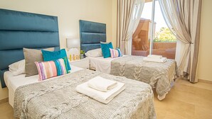 Une chambre d’hôtel avec deux lits, chacun recouvert d’une couverture tricotée et agrémenté d’oreillers, une table de chevet avec une lampe, et une vue sur l’extérieur par une fenêtre munie de rideaux.