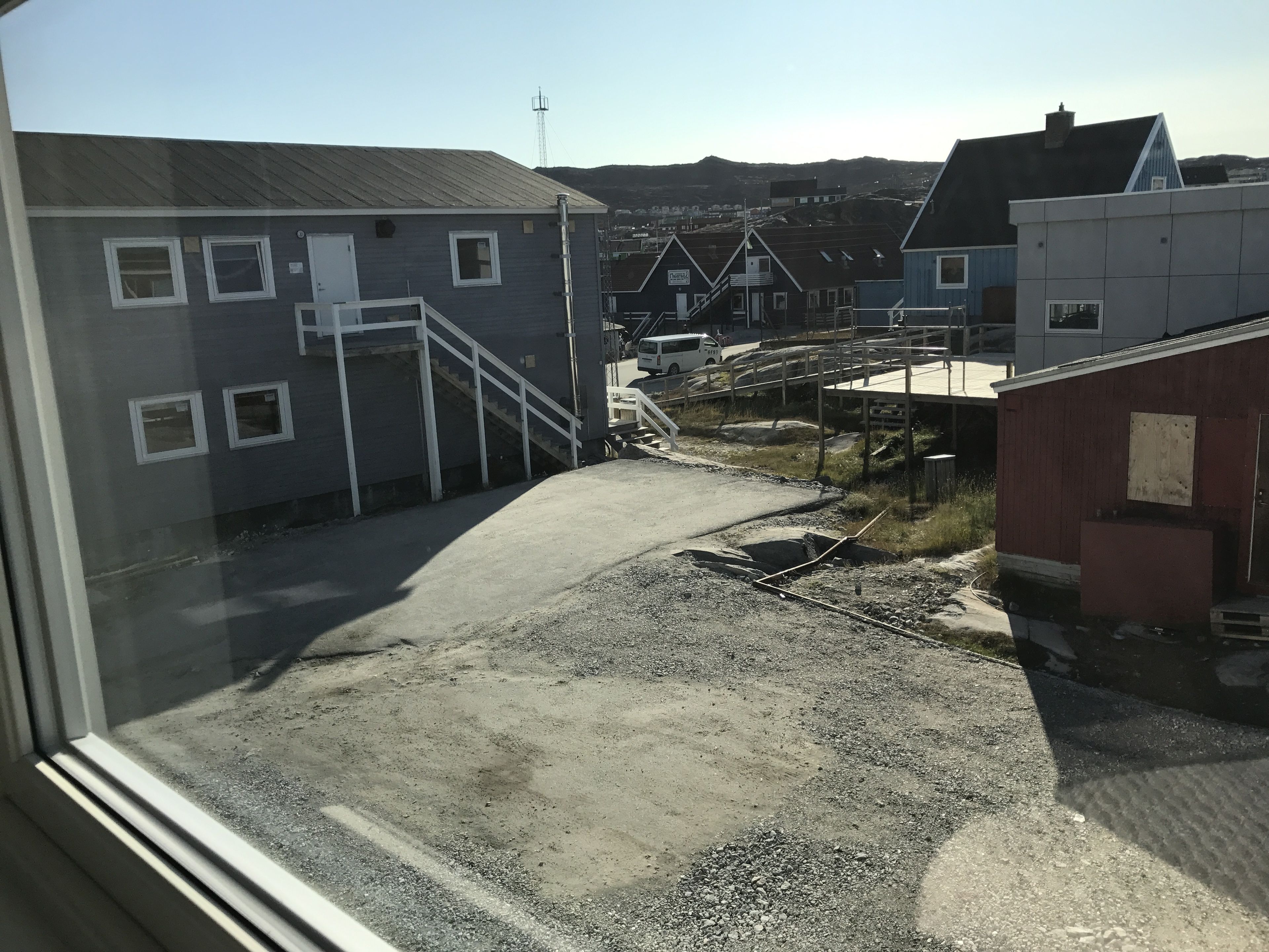 On voit un bâtiment avec un escalier menant à une porte, une zone recouverte de gravier et, au premier plan, un bâtiment rouge.