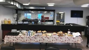 A buffet setup with various food items and beverages on a table, a kitchen area in the background, and a television mounted on the wall. - Hotel Nuka (Ilulissat)