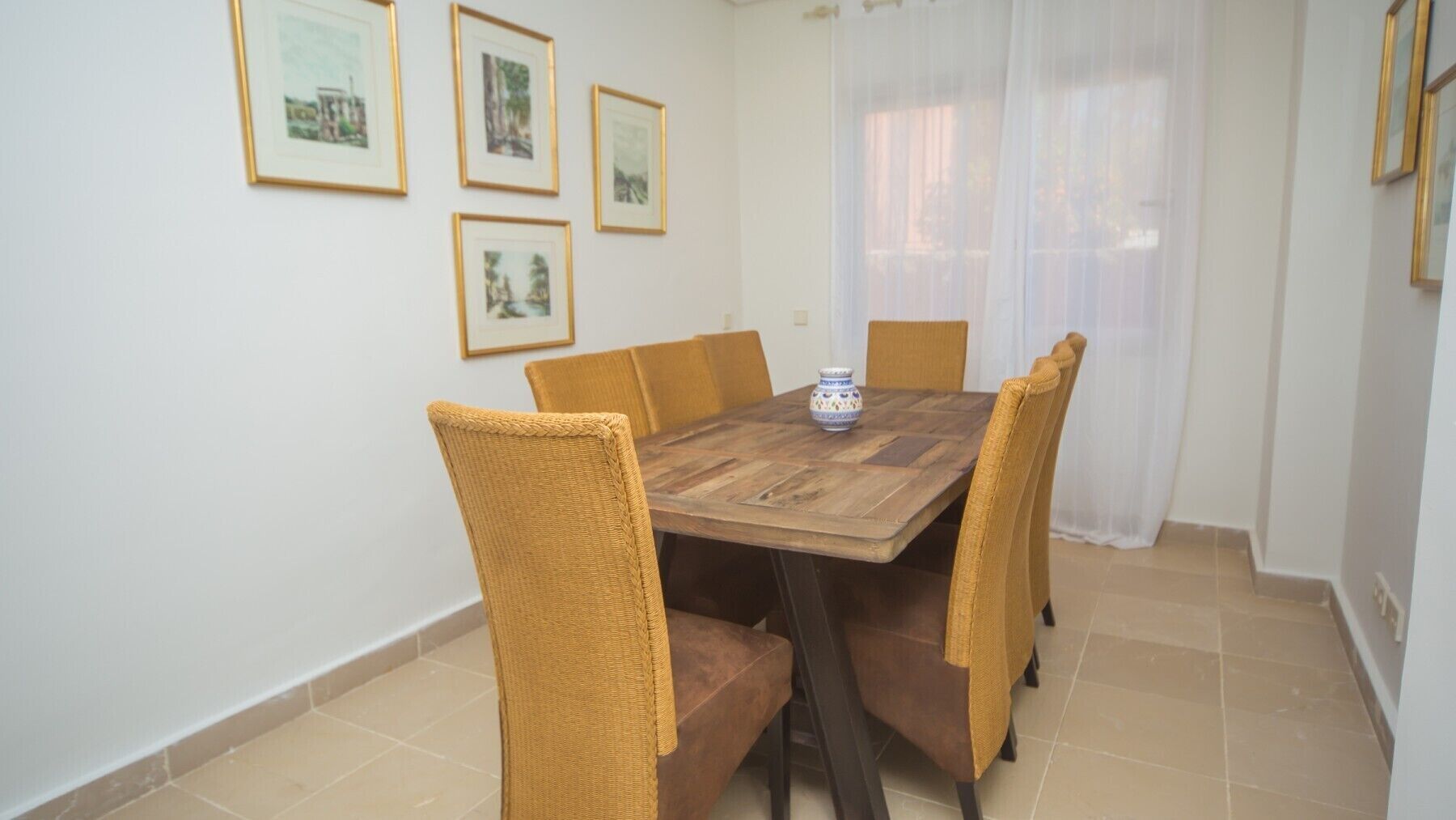 A dining area with a wooden table, six wicker chairs, and four framed pictures on the wall.