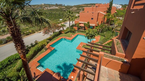 En swimmingpool med træterrasse, omgivet af grønt og bygninger med terracotta tagsten.