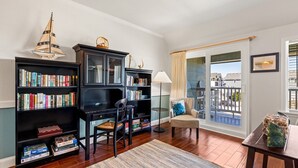 Un salon doté d’un plancher en bois, d’une bibliothèque, d’un bureau, d’une chaise, d’une lampe, d’une fenêtre donnant sur un beau paysage et d’un balcon.