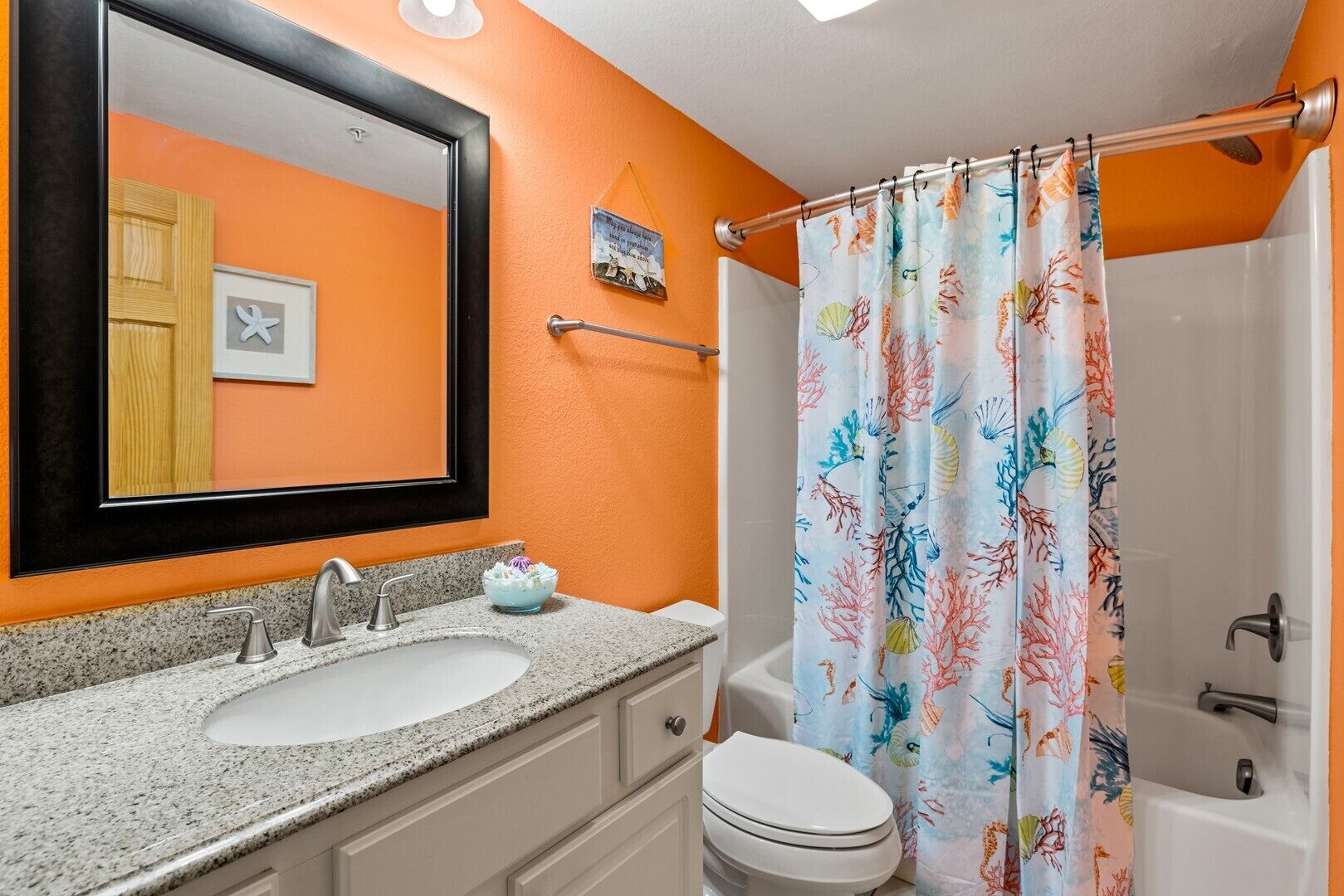 Une salle de bain avec un mur orange, un lavabo blanc, un miroir, des toilettes et un rideau de douche orné d’un motif de récif corallien.