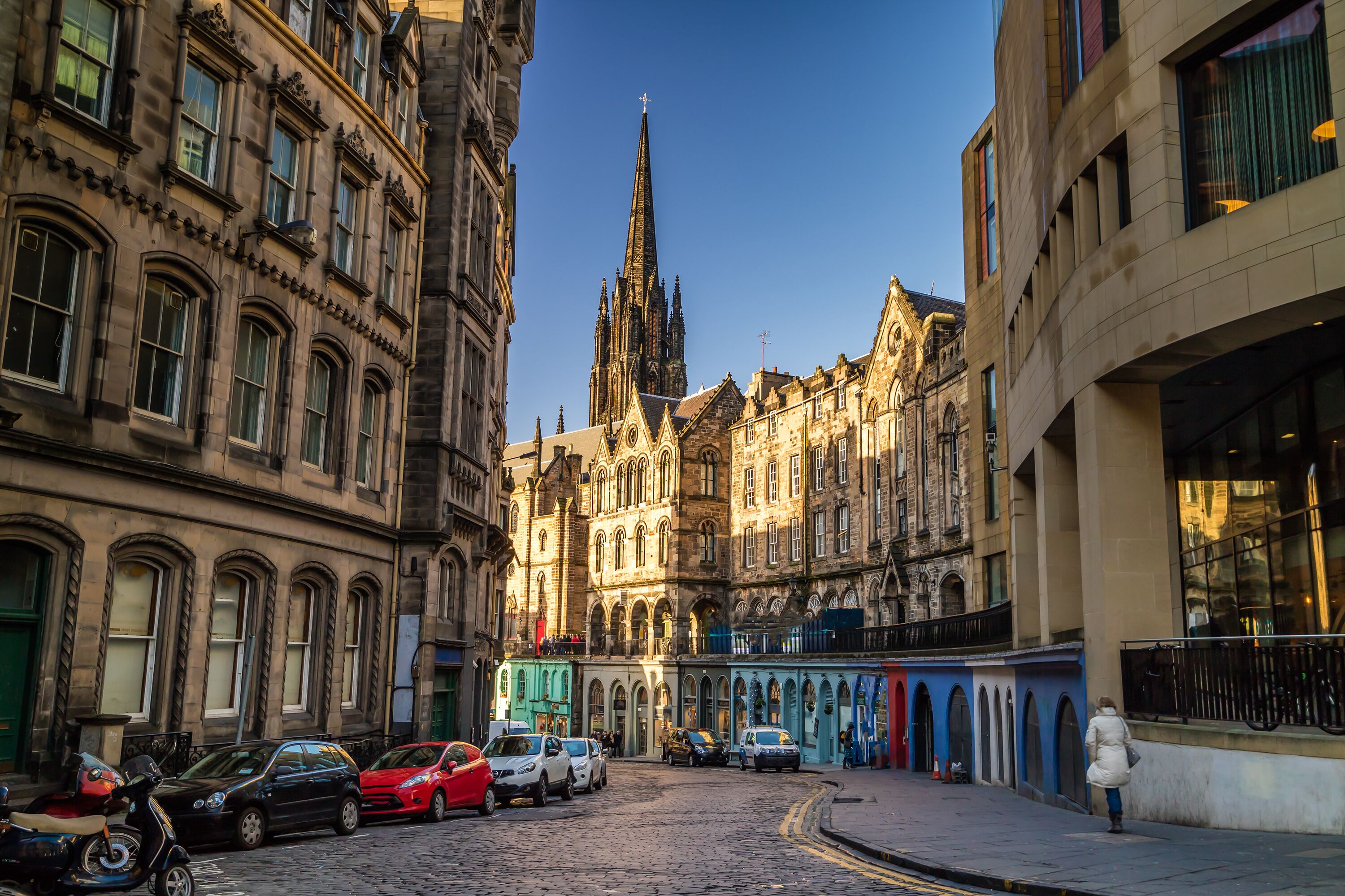 A cobblestone street with historic buildings, a church spire, and parked cars.