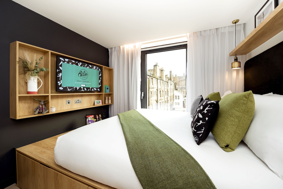 A hotel room with a bed, a mounted TV, a window with curtains, and a city view.