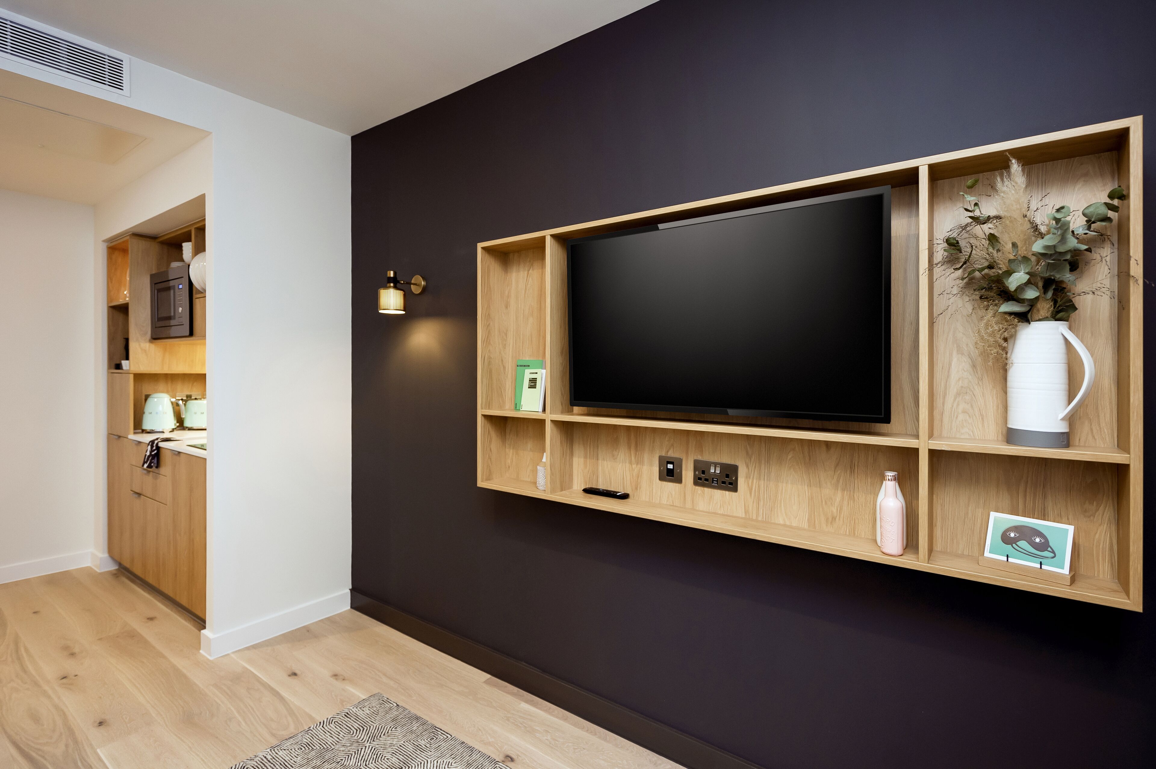 A modern living room with a large flat-screen TV mounted on a wooden wall unit, a kitchen area with wooden cabinets, and a decorative vase with dried plants.
